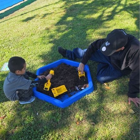 a parent and his son play with toy trucks in a small sensory bin with dirt