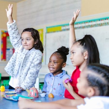 four girls are in a classroom and two of them are raising their hands