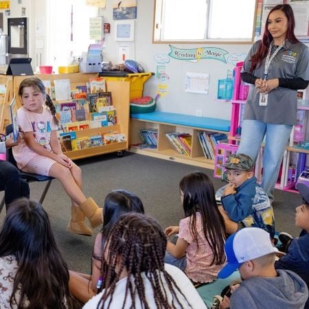 a classroom with a young girl sitting in a chair in front of a group of students sitting on the floor with an Options for Learning teacher standing up watching the children