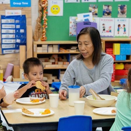 teacher sitting at a table of preschool-age children eating