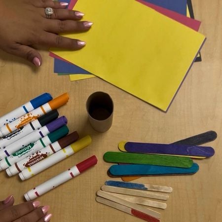 table with colored construction paper, an empty toilet paper roll, colored popsicle sticks, and markers