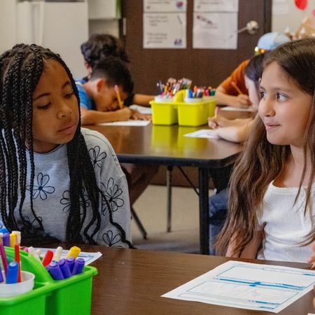 two school-age children sit at a desk and work on a worksheet together in school