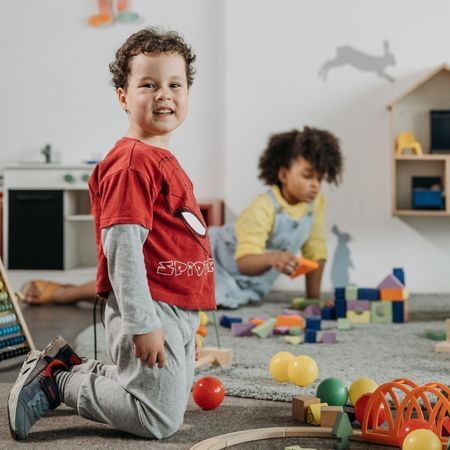 a young boy in a Spider-Man t-shirt plays with blocks on the rug of a classroom and smiles at the camera