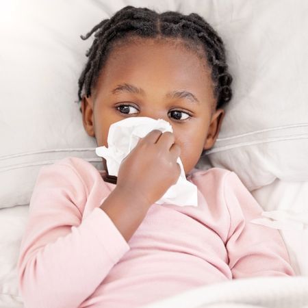a young girl blows her nose into a tissue while laying down and wearing a pink long sleeve shirt