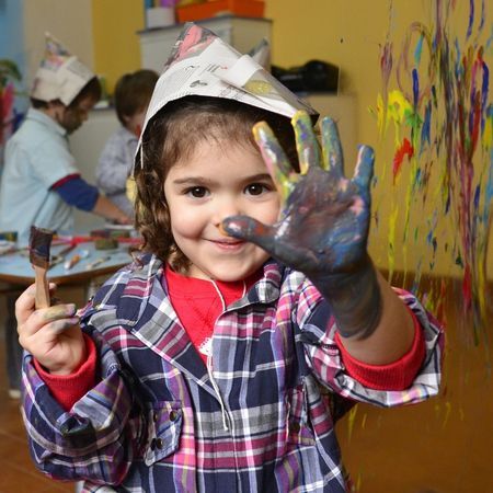 a young girl smiles at the camera wearing a hat made out of newspaper and shows her hand painted in paint