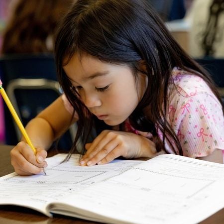 a young girl writing in a notebook with a pencil at a desk