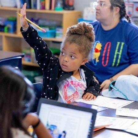 a young girl raising her hand in a classroom while sitting at a desk