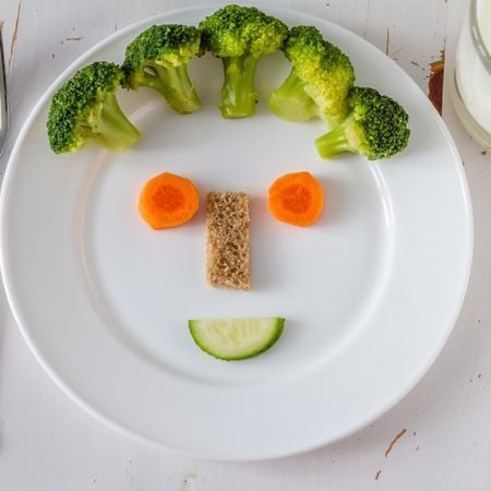 a plate with a smiley face made with broccoli, carrots, a piece of bread and a cucumber