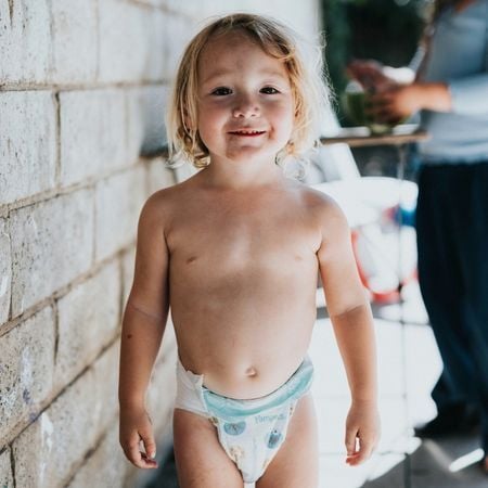 a toddler-age boy stands in his diaper and smiles at the camera