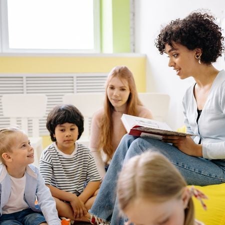 a group of students (2 girls and a boy) listen to a female teacher reading