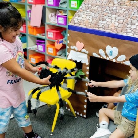 two preschool age girls are in a classroom holding both sides of a stuffed spider