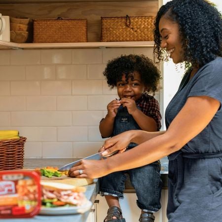 a young boy sitting on the countertop while his mom is cutting up vegetables and making a sandwich